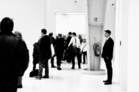 „Ein Angestellter steht reglos im Ausstellungsraum, während sich Besucher:innen um ihn herum bewegen und unterhalten. Momentaufnahme des Alltags.