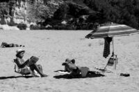 Ein Mensch sitzt am Strand, mit Hut, in einem Stuhl lesend. Momentaufnahme des Alltags.