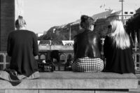 Drei Frauen auf einer Steinbank, Rückenansicht – zwei im Gespräch, eine vornübergebeugt, vertieft ins Handy. Momentaufnahme des Alltagsgeschehens.“